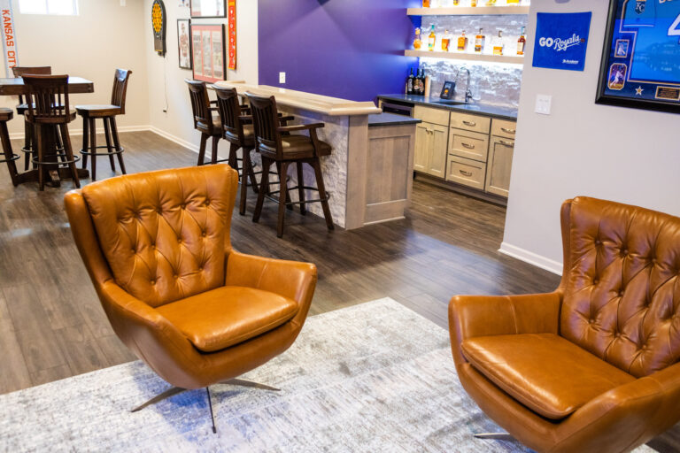 A home bar with underlit shelves for alcohol near stools and leather lounge chairs after basement remodeling in Leawood